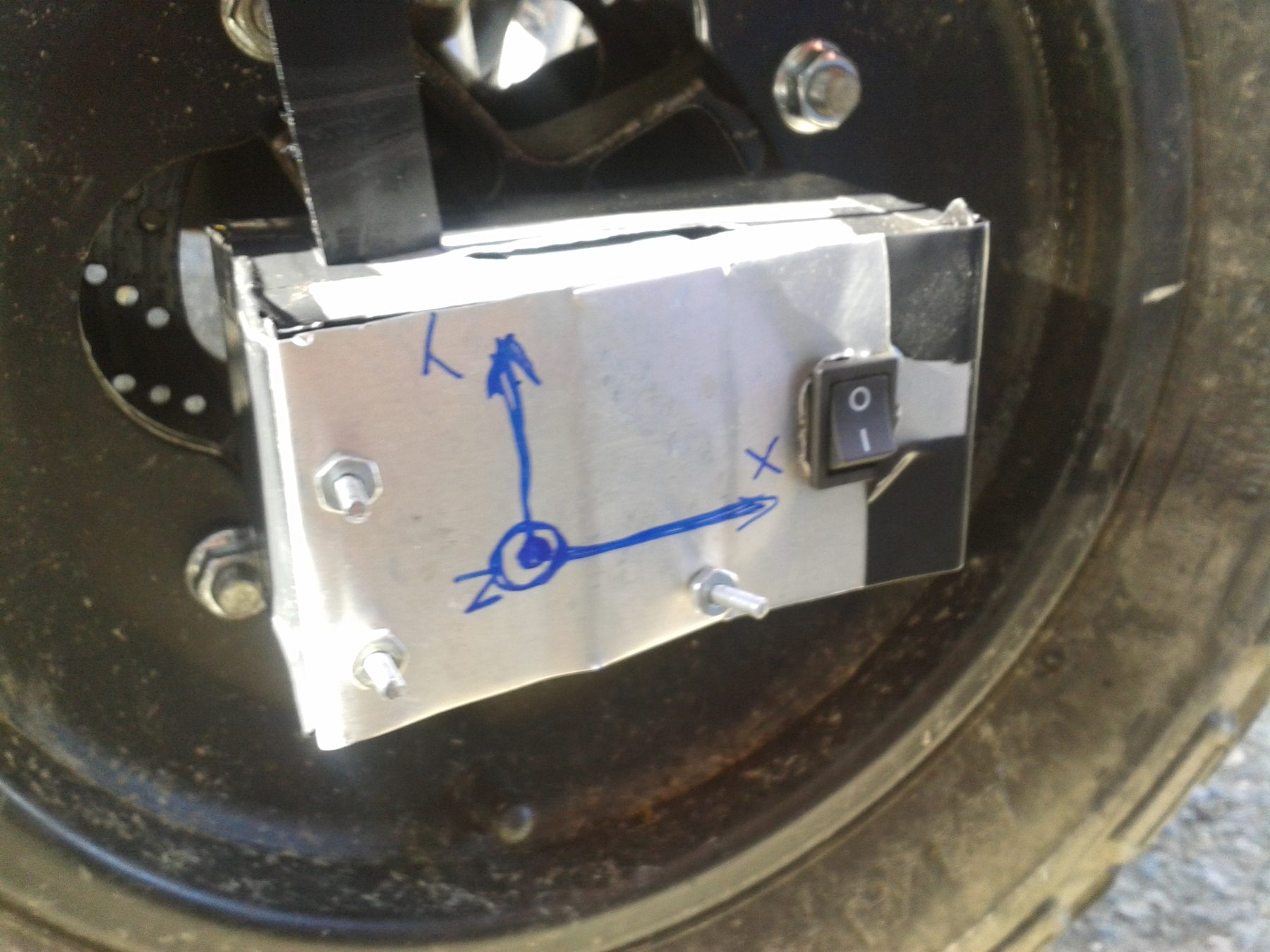 A hand-formed aluminium enclosure bolted to a Baja SAE USB wheel hub, with X, Y and Z axes hand-drawn on it in blue marker and a power switch on the side
