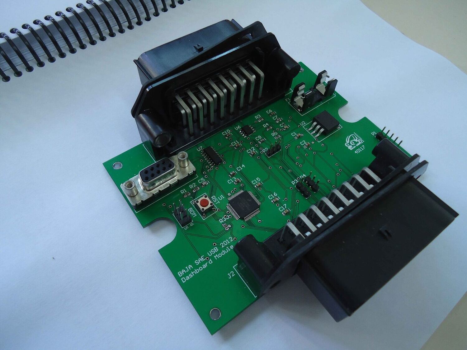 The assembled dashboard-module board, silkscreened "BAJA SAE USB 2012 &mdash; Dashboard Module": a 64-pin microcontroller in the centre, a DB9 and reset button at left, and large keyed connector housings plugged into the two harness headers