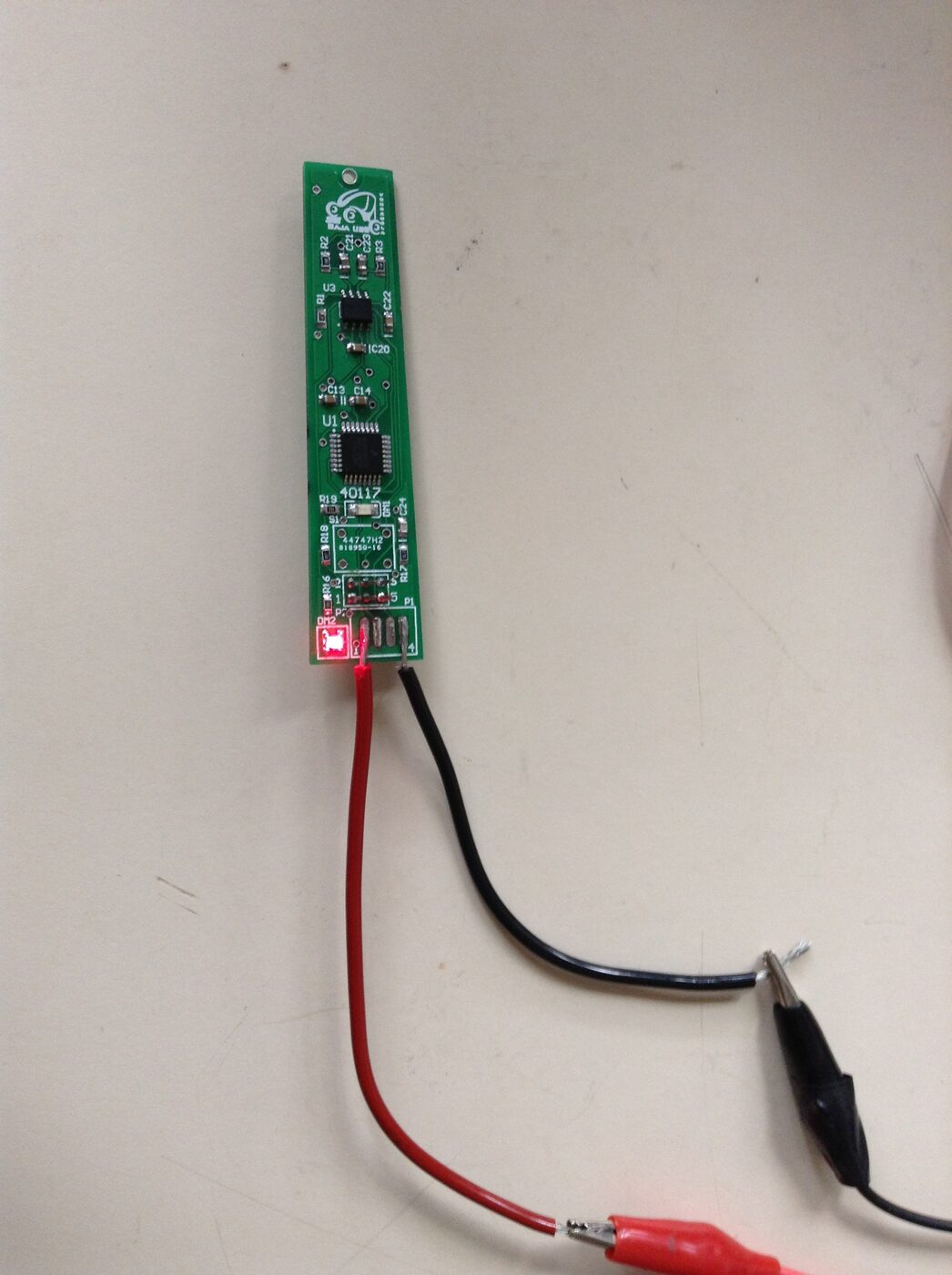 The assembled fuel-level module: a long, narrow green PCB with a Baja USB rabbit logo, powered on the bench from a lab supply through red and black clip leads, its power LED lit