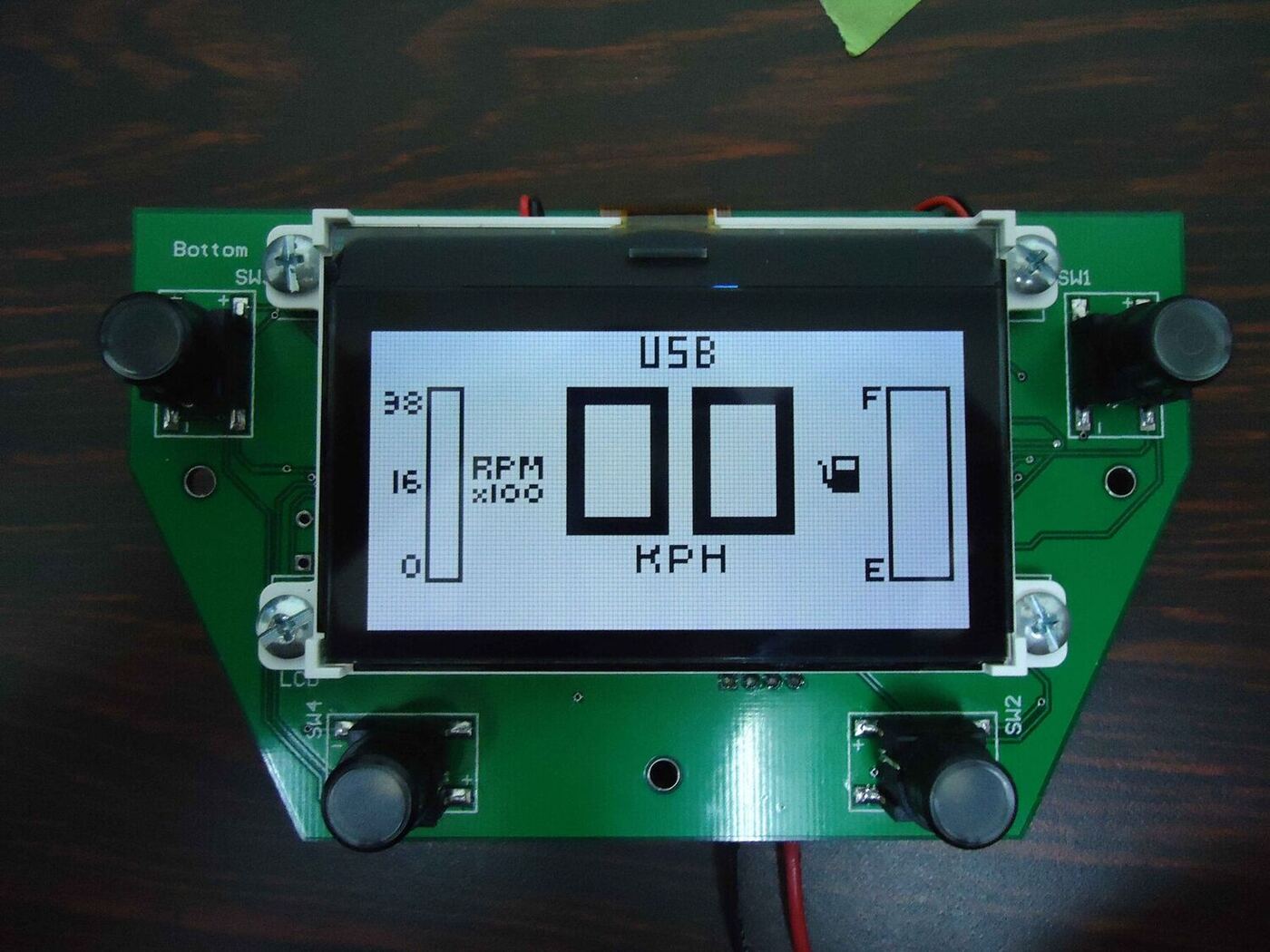 The steering-wheel board running on the bench: a chevron-shaped PCB with an illuminated button at each corner and a graphic LCD in the centre showing "USB", a large "00 KPH" speed readout, an RPM bar down the left and a fuel bar down the right