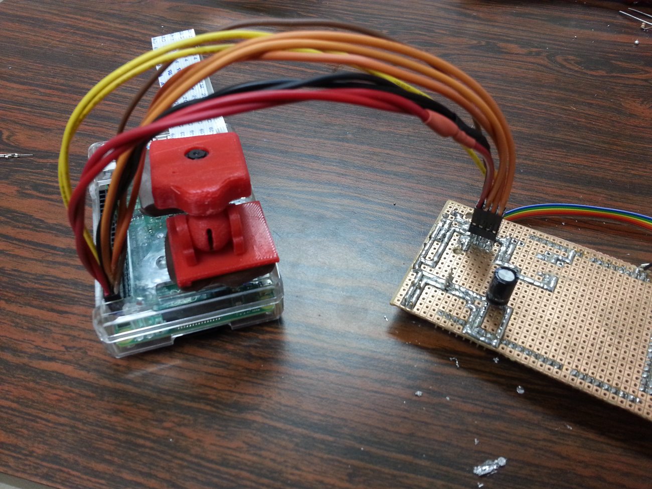 A Raspberry Pi in a case with a red 3D-printed pan-tilt camera mount, connected by ribbon cable to the Arduino shield perfboard