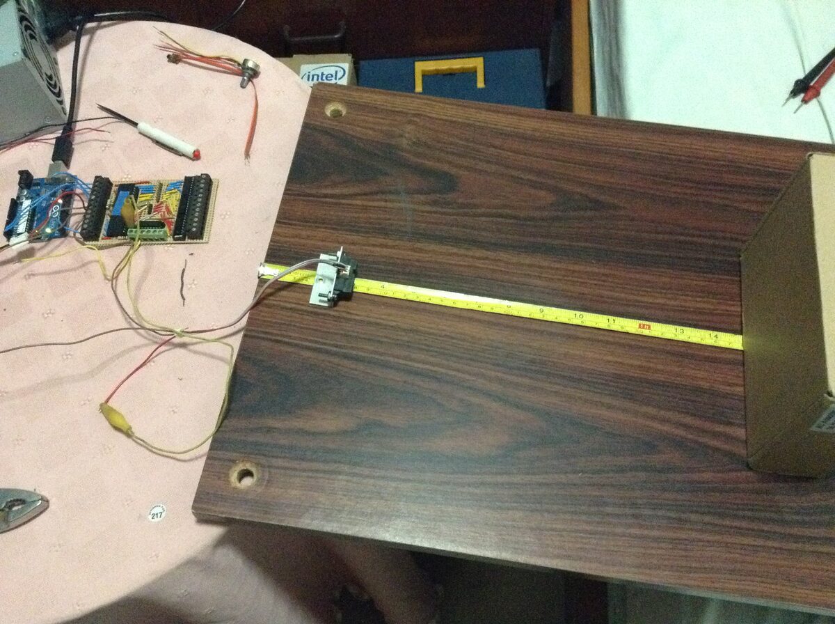 The Sharp IR sensor clamped on a small bracket at the zero end of a yellow tape measure on a wooden board, aimed at a cardboard target, with its wires running back to an Arduino Uno and a PIC perfboard with terminal blocks